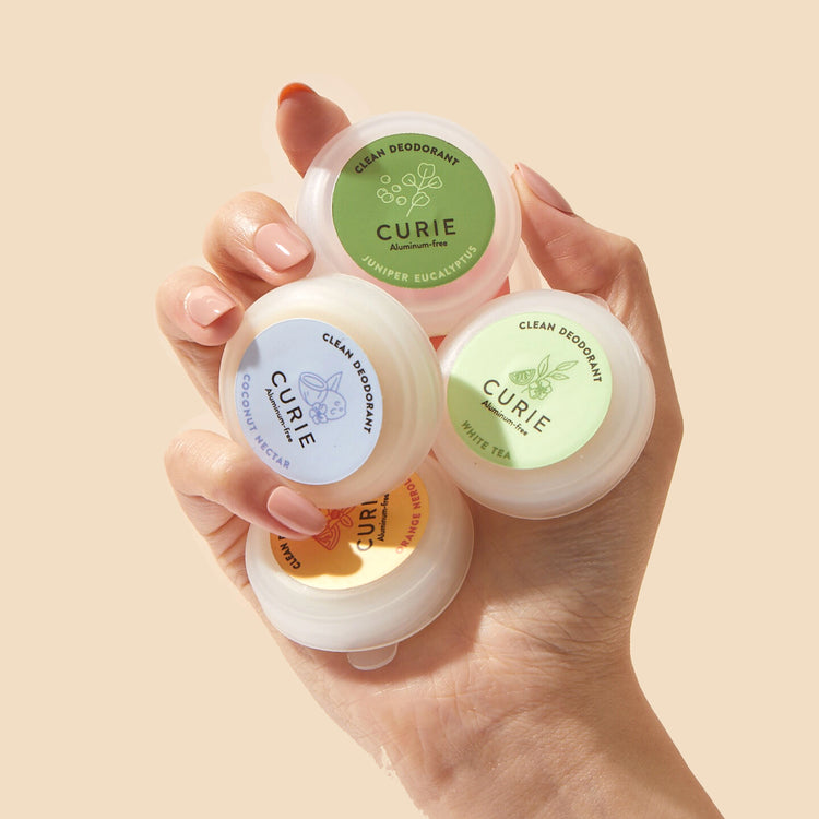 A hand holds four different scents of Curie clean deodorant in small containers.